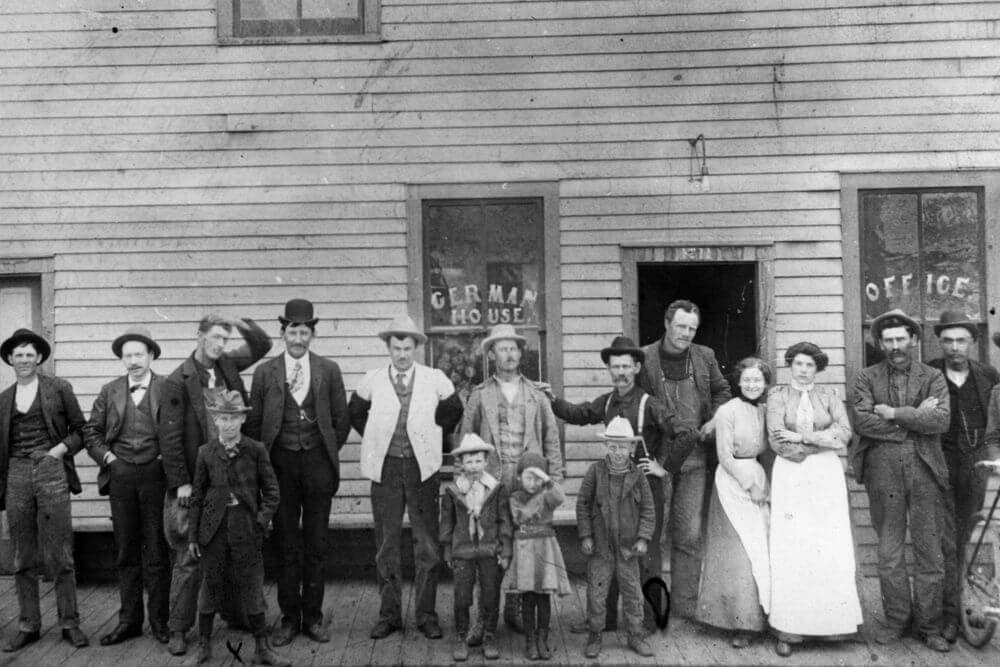 A large group of men, women, and children pose together outside a wooden building with signs reading "German House" and "Office."