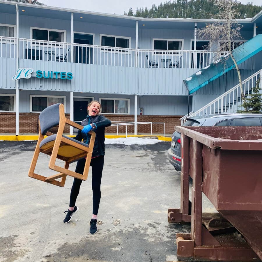 A woman joyfully lifts a chair in front of a motel, with a dumpster and parked cars nearby.