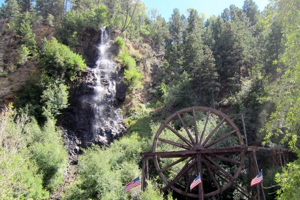 A waterfall flows down a rocky hillside beside a large wooden water wheel, surrounded by trees and American flags.