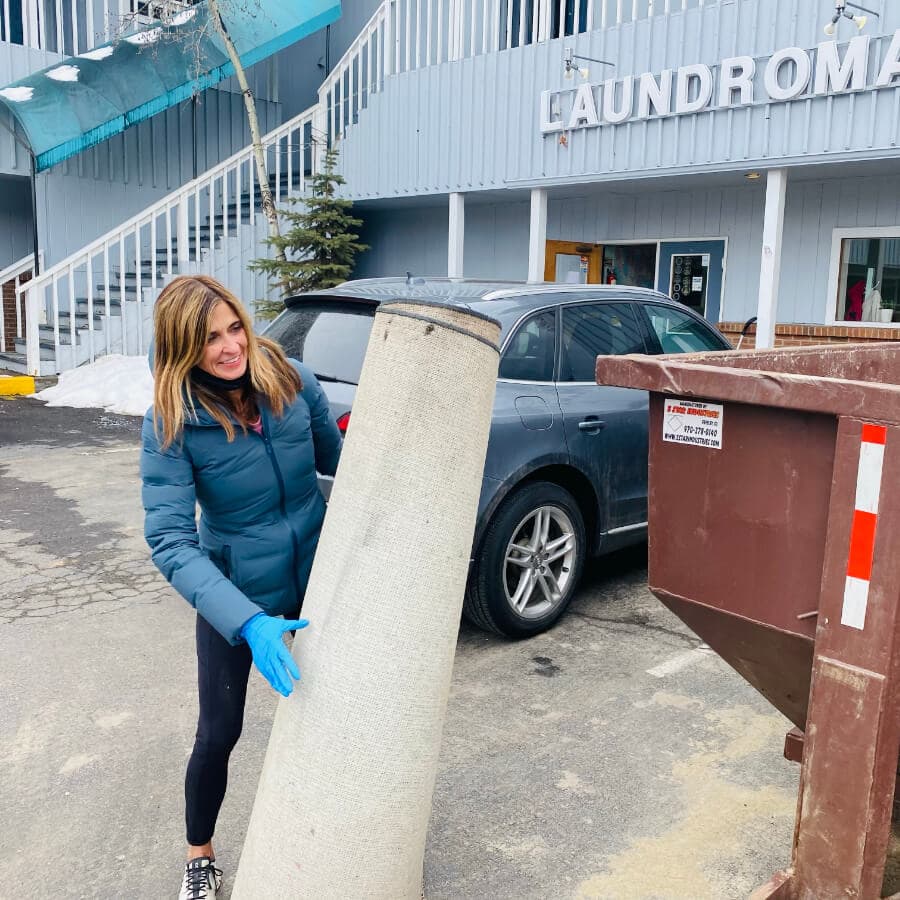 A woman in a blue jacket is lifting a rolled carpet towards a dumpster outside a laundromat.