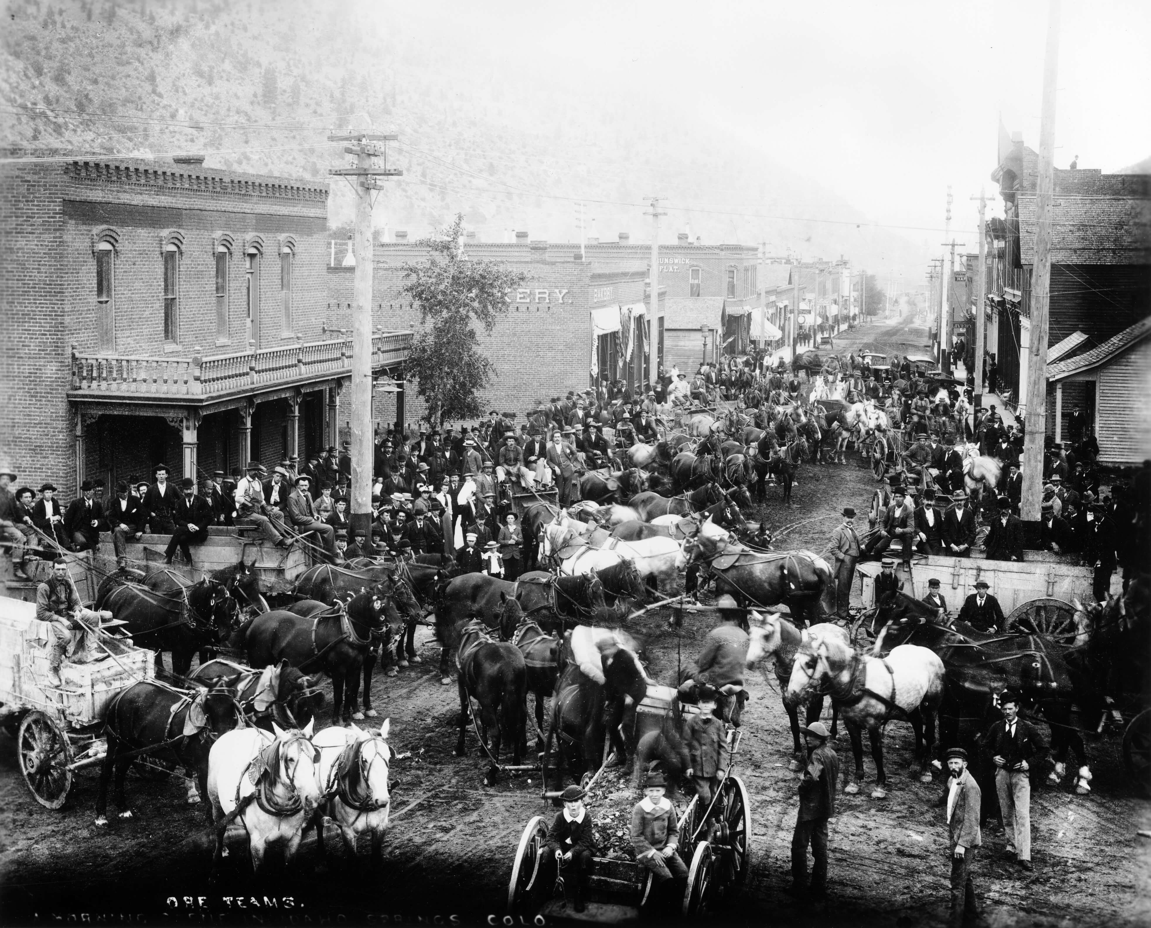 A bustling historical street scene filled with horse-drawn wagons and people in early 20th-century attire.