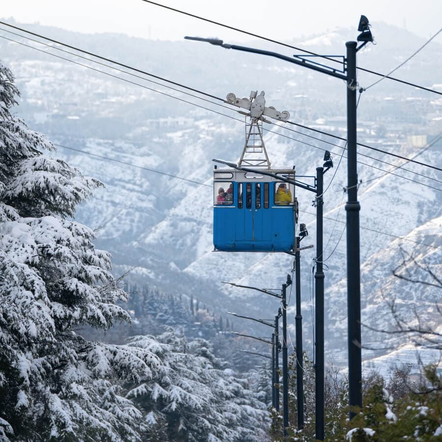 A blue cable car ascends over snow-covered mountains.