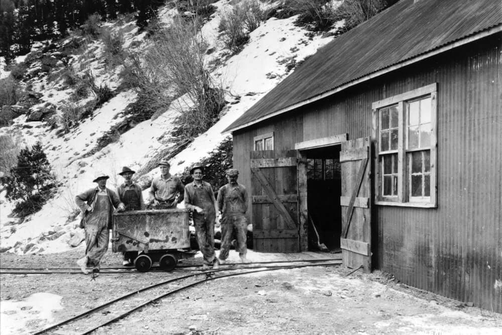 A group of five miners stands outside a wooden mining building, with a mine cart on tracks in front of them.